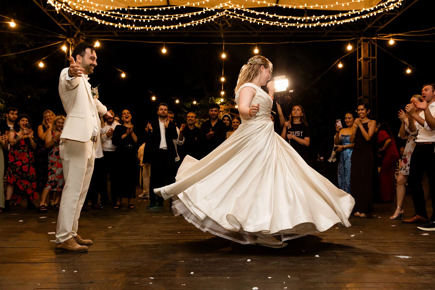 A joyful bride spins on a dance floor under string lights as her groom and guests cheer during a Turkish-American Sarıyer Korusu wedding.