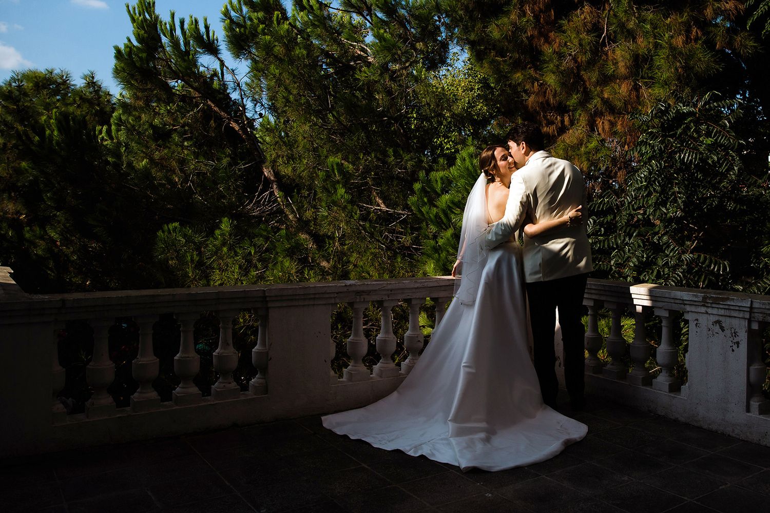 A romantic portrait of a bride and groom embracing on a stone balcony, dramatically lit by golden hour sun with pine trees behind them.