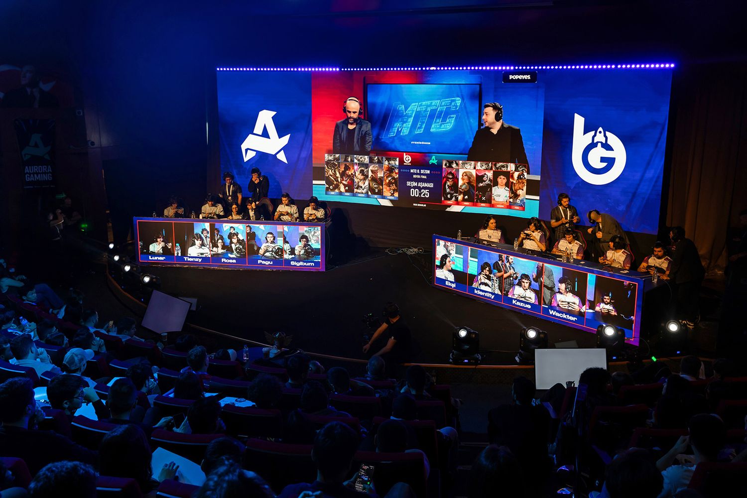 A wide view of an auditorium filled with attendees watching the player draft at a live esports gaming tournament on a massive stage screen.