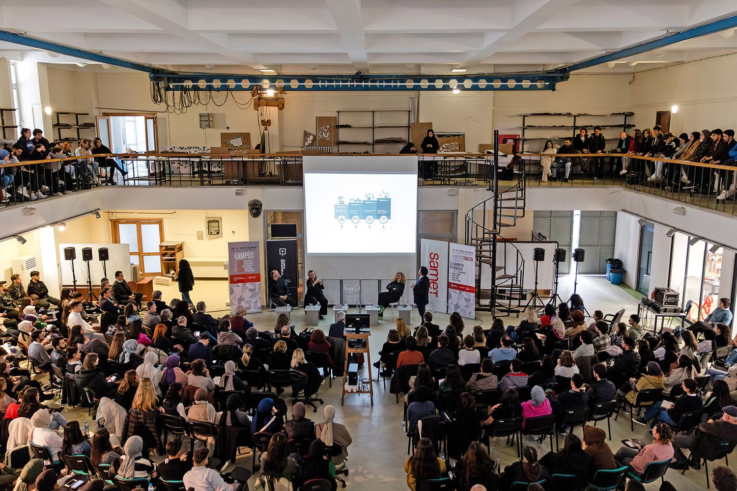 A wide, elevated view of a large audience filling an expansive industrial-style venue to watch a corporate design panel presentation.