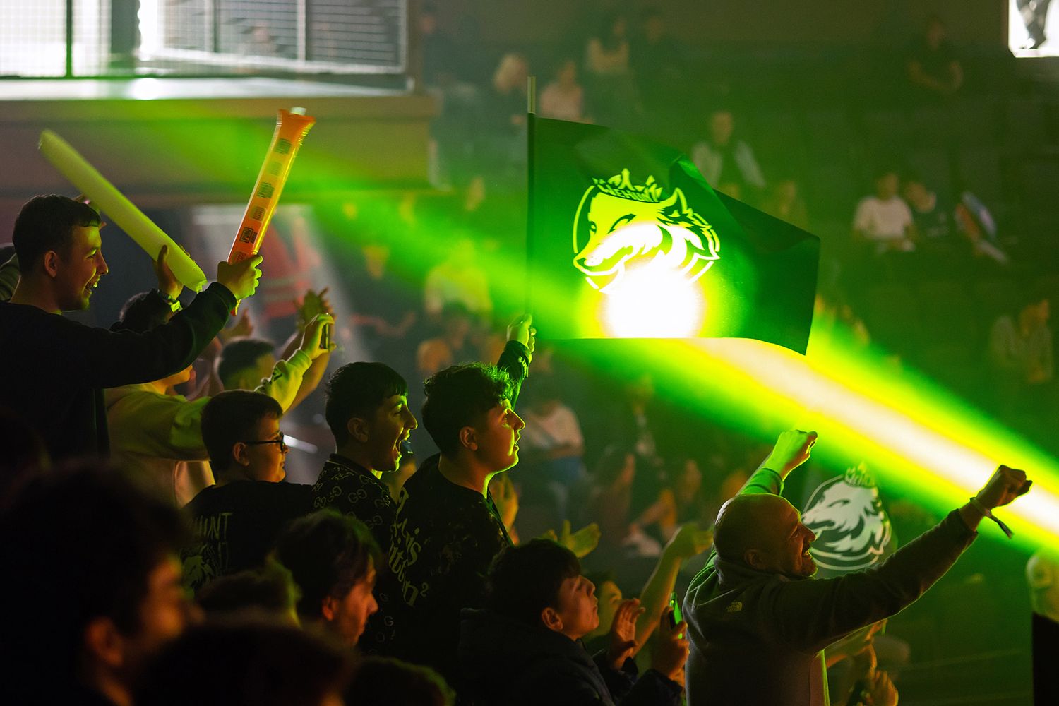 Passionate esports fans cheering and waving brightly lit green team flags in a dark, atmospheric tournament arena.