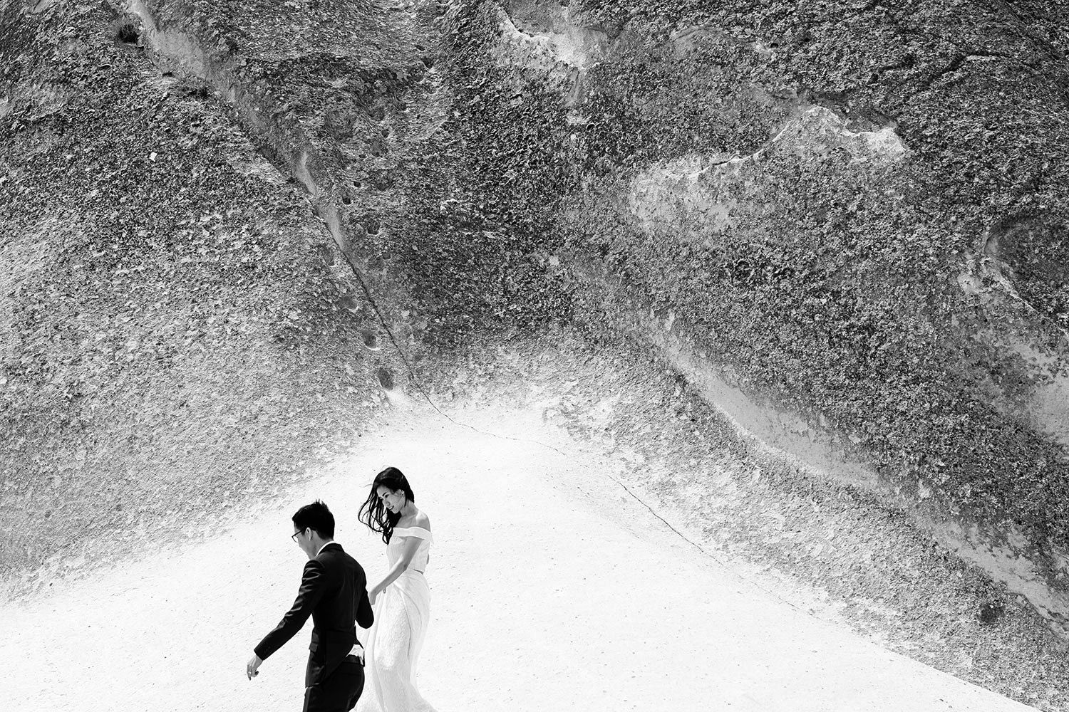 A black and white documentary-style portrait of a bride and groom in wedding attire walking together across the massive, textured rock forma