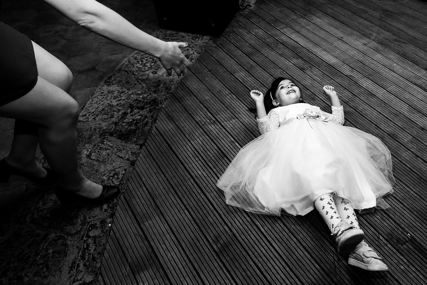 Candid black and white photo of a laughing flower girl playfully lying on a wooden deck at an Elio Sedef Island wedding.