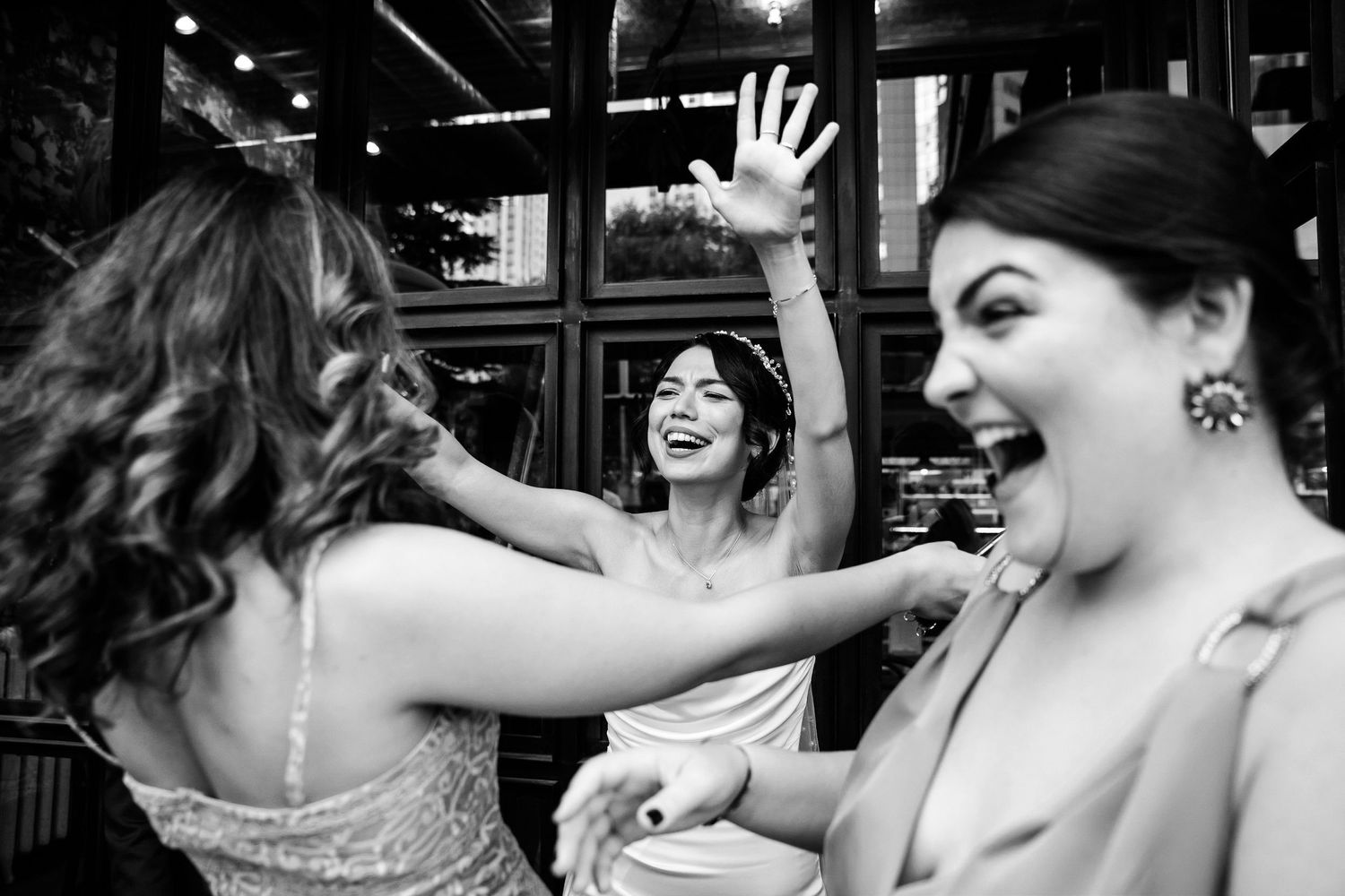 Black and white candid photo of a joyful bride with her arms raised, laughing enthusiastically with her bridesmaids on her wedding day.