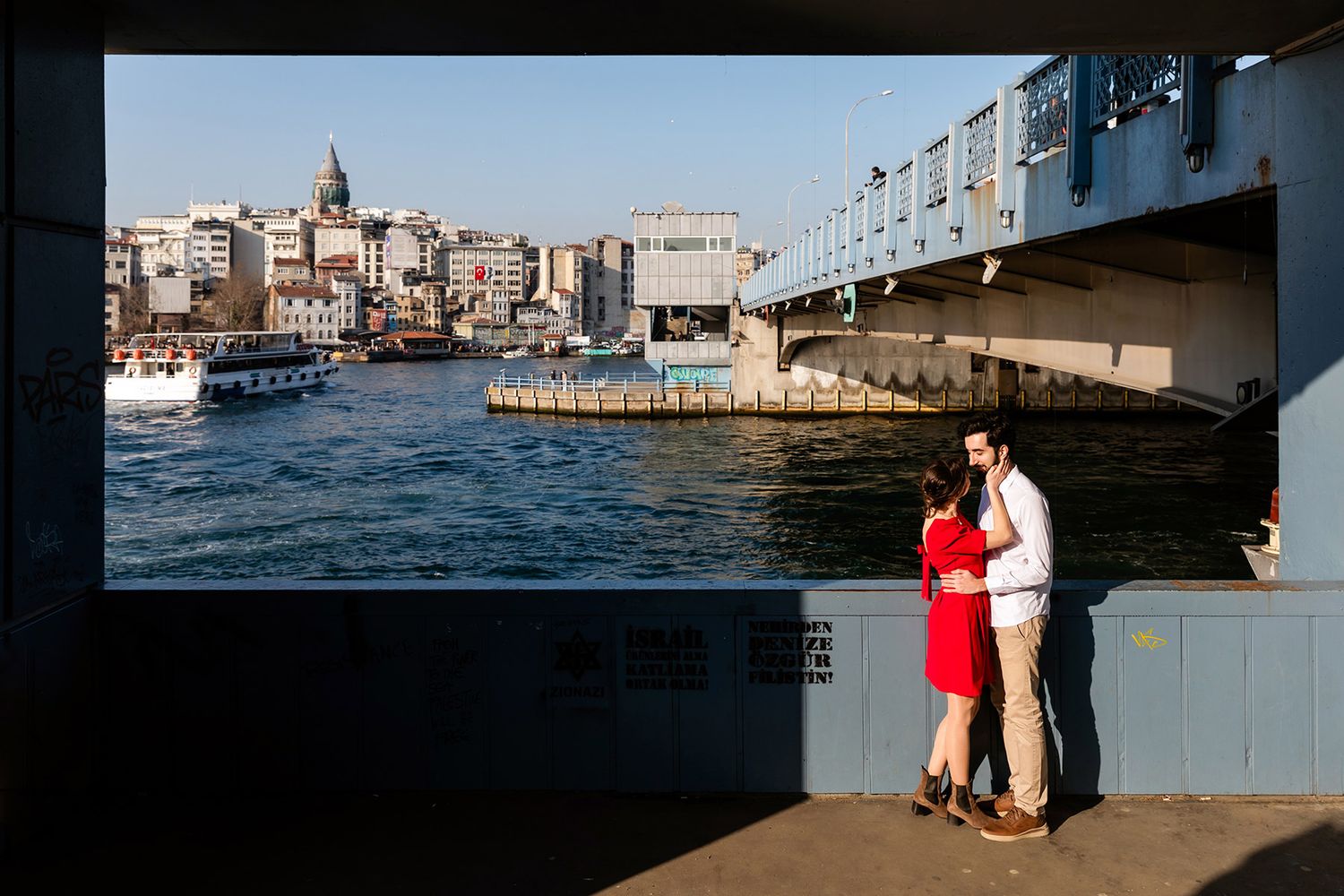 A couple in red and white embraces beneath the Galata Bridge, perfectly framed with the Galata Tower and Bosphorus water in the background.