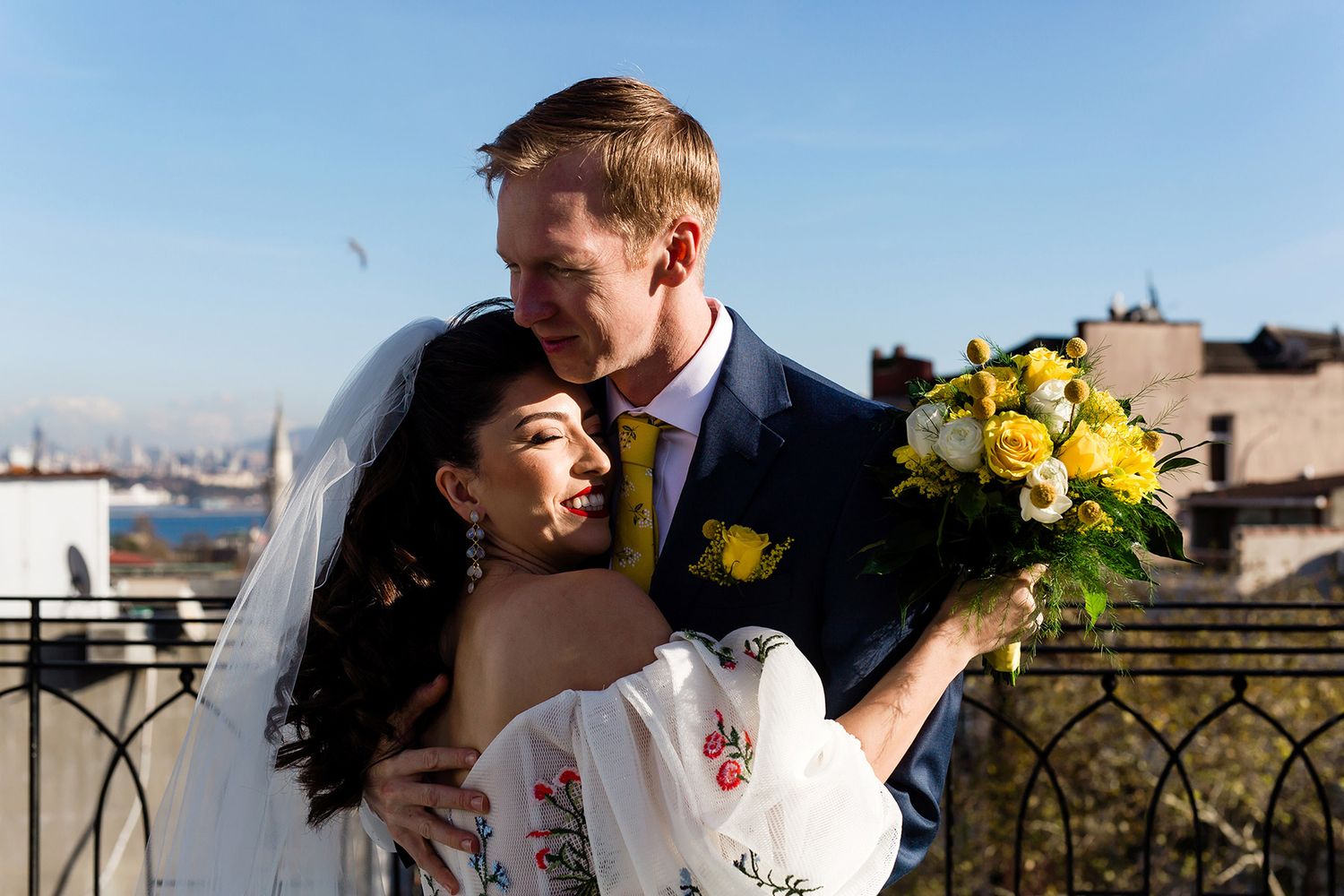 A joyful bride with a yellow bouquet hugs her groom during a first look on an Istanbul rooftop, with the city skyline in the background.