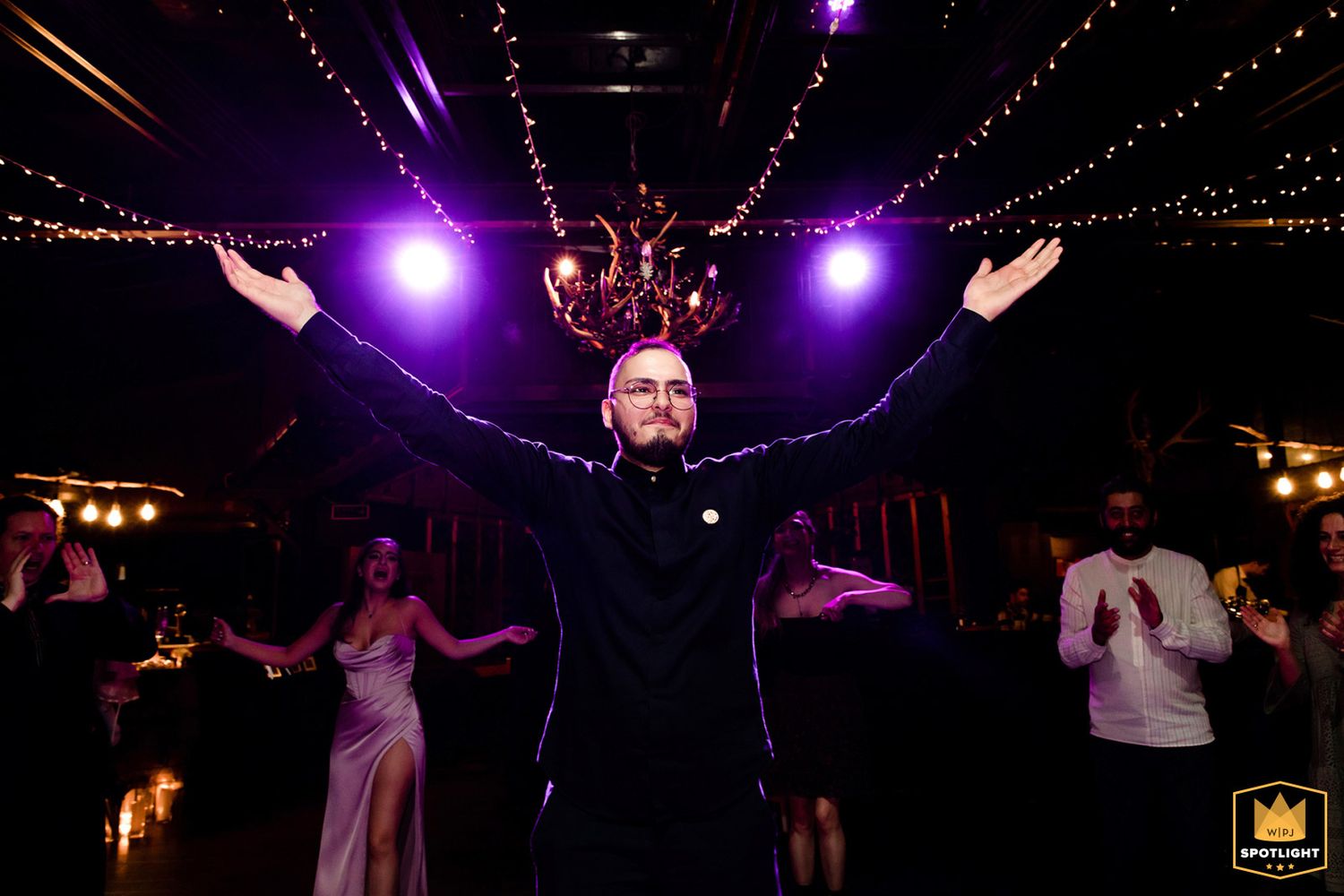 WPJA award-winning candid reception photo of a man standing on the dance floor with arms raised wide under vibrant purple party lights.