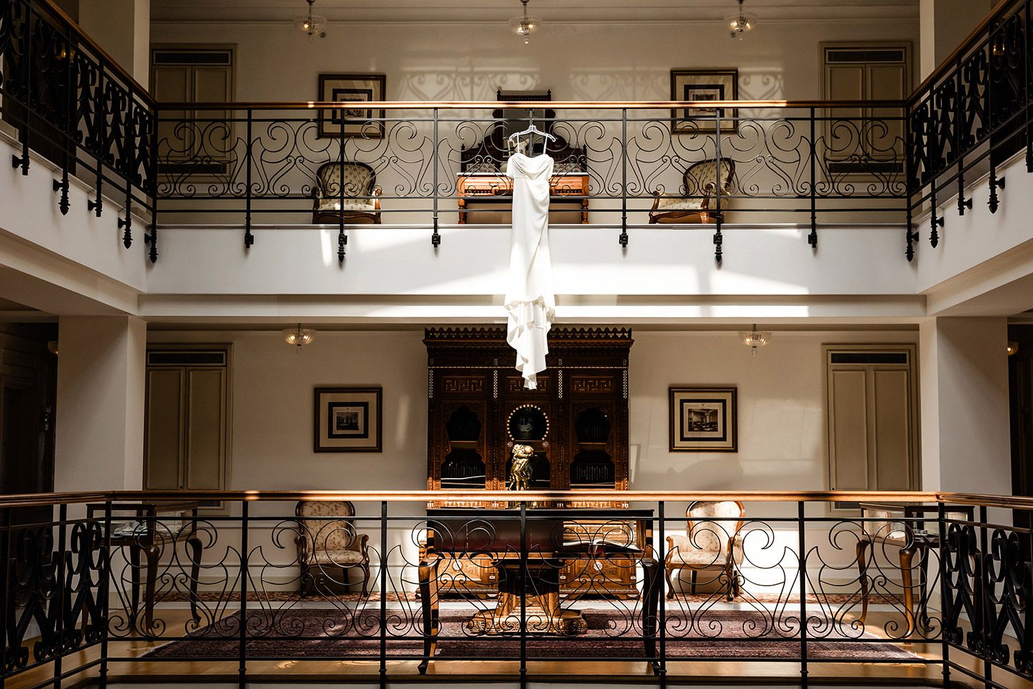 An elegant white wedding dress hangs from an ornate iron balcony in the historic, luxurious atrium of the Pera Palace Hotel in Istanbul.