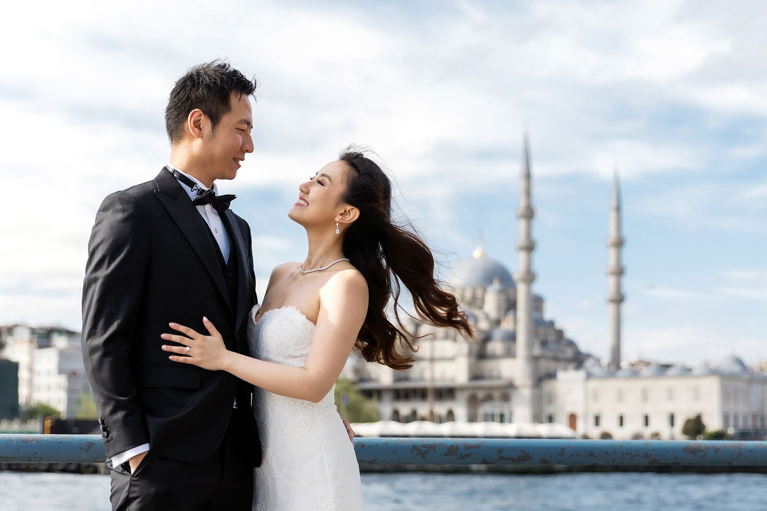 An elegant bride and groom embrace on the Galata Bridge, wind blowing her hair, with Istanbul's iconic Yeni Cami mosque in the background.