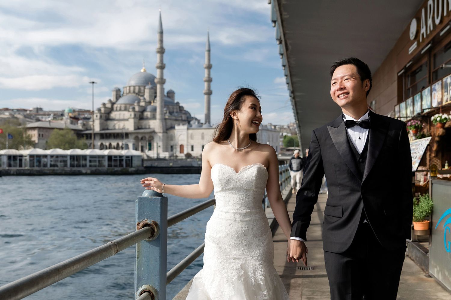 A happy couple in wedding attire walks hand-in-hand along the Galata Bridge with the historic Süleymaniye Mosque behind them in Istanbul.