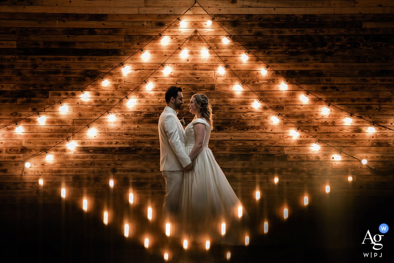 AG WPJA award-winning creative night portrait of a couple standing in front of a wooden wall illuminated by geometric string lights.