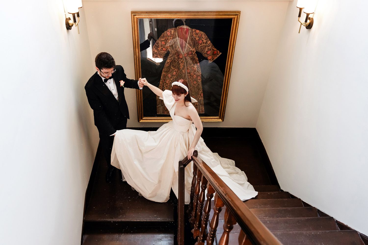 A groom holds his bride's hand as they walk down a wooden staircase past an antique garment at Esma Sultan in Istanbul.