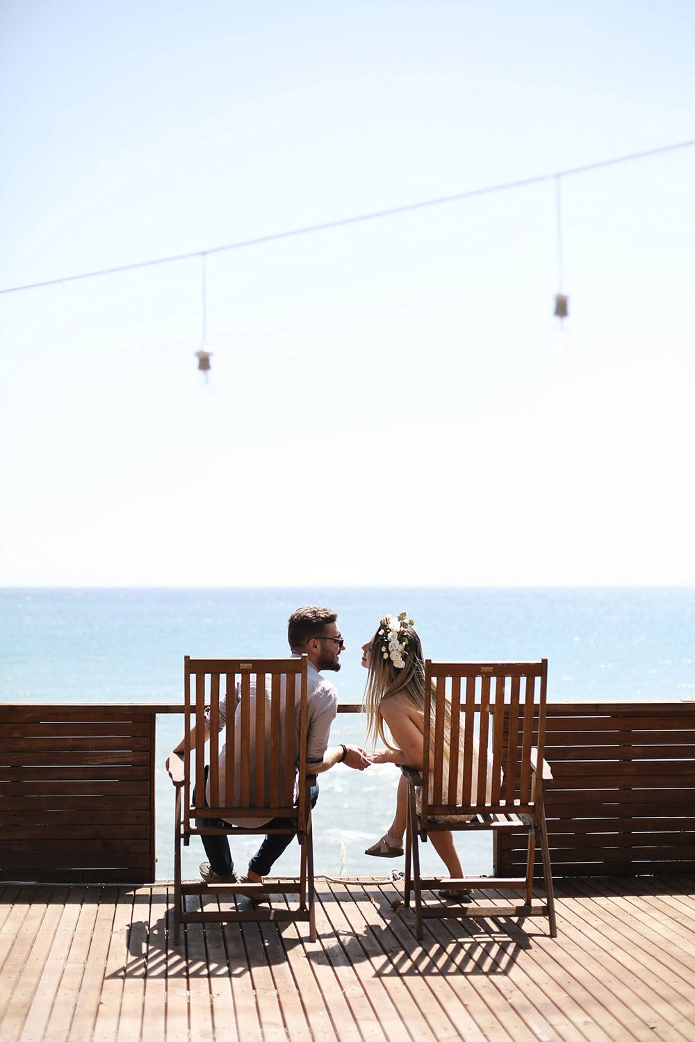 An engaged couple sits in wooden chairs on a sunlit pier, holding hands and looking at each other with the blue sea of Tekirdağ behind them.