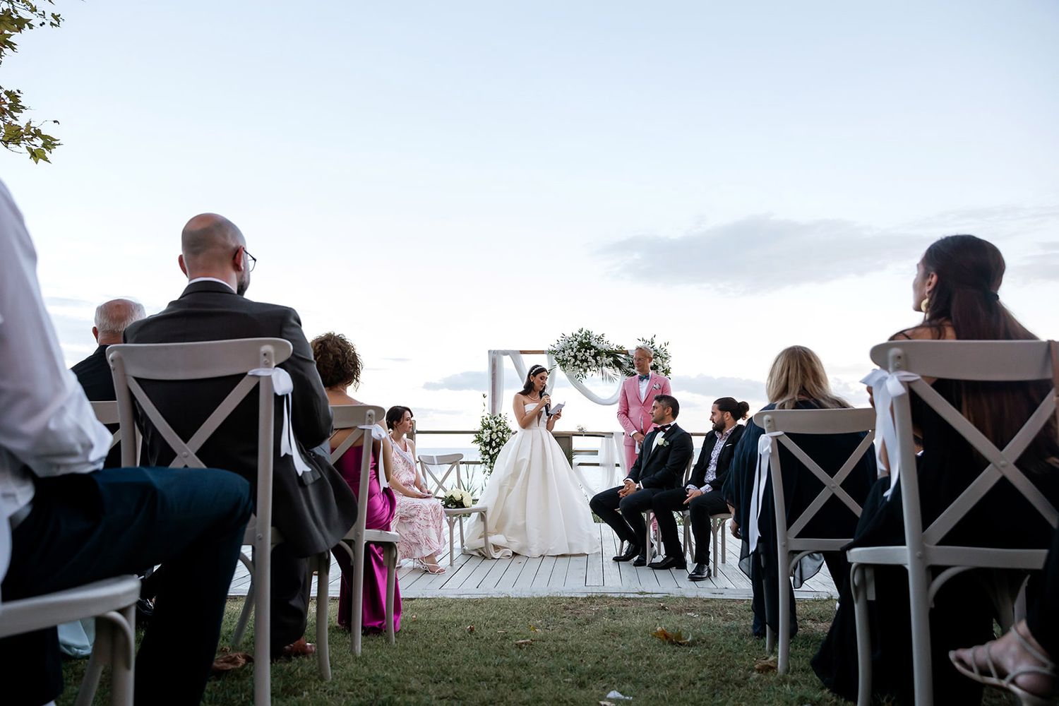 A wider view of an outdoor sunset wedding ceremony, showing guests in cross-back chairs watching the bride read vows at the altar.