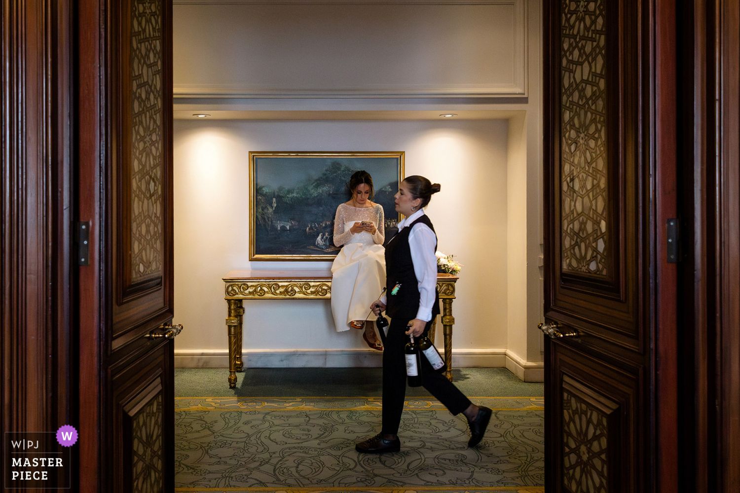 WPJA award-winning documentary photo of a bride checking her phone on a hallway as a server walks by with wine in Istanbul Ciragan Palace