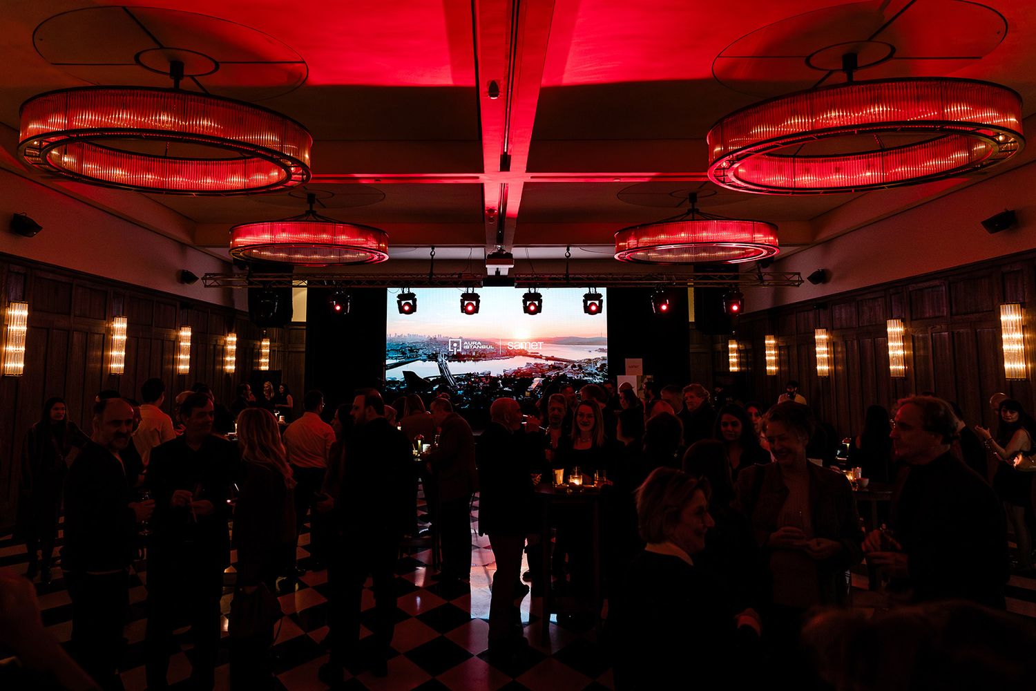 A wide view of a stylish corporate networking gala featuring dramatic red chandeliers, a checkered floor, and an Aura Istanbul stage screen.