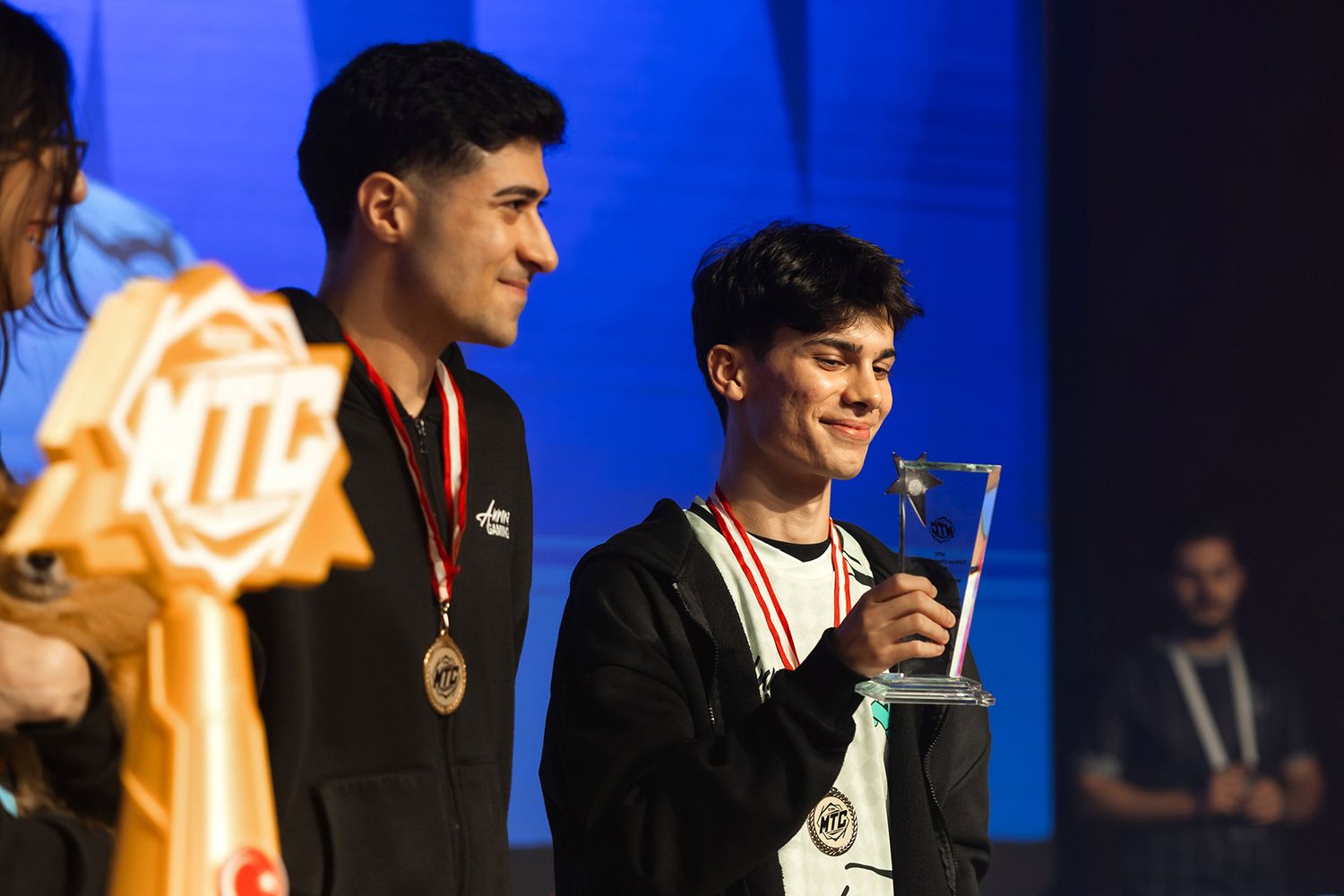 Two smiling esports players proudly holding a glass championship trophy and medals on stage after winning a major gaming tournament.