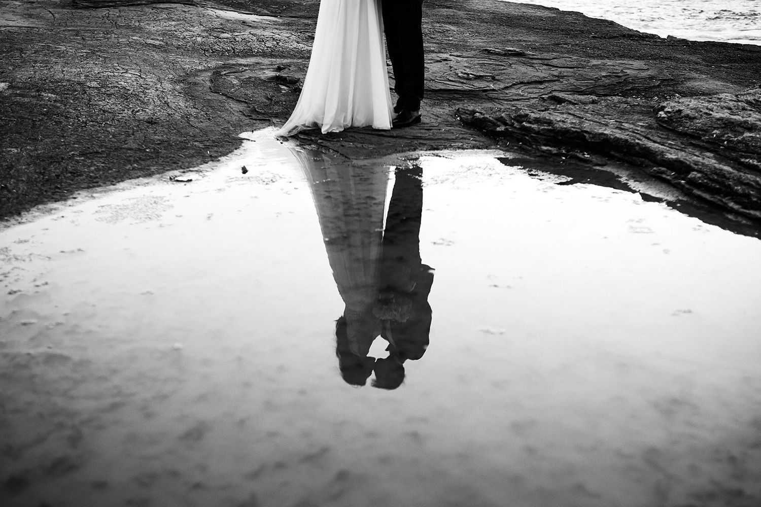 An artistic black and white reflection of a bride and groom kissing, captured in a water puddle on the rugged rocky coast of Kizil Kayalar.