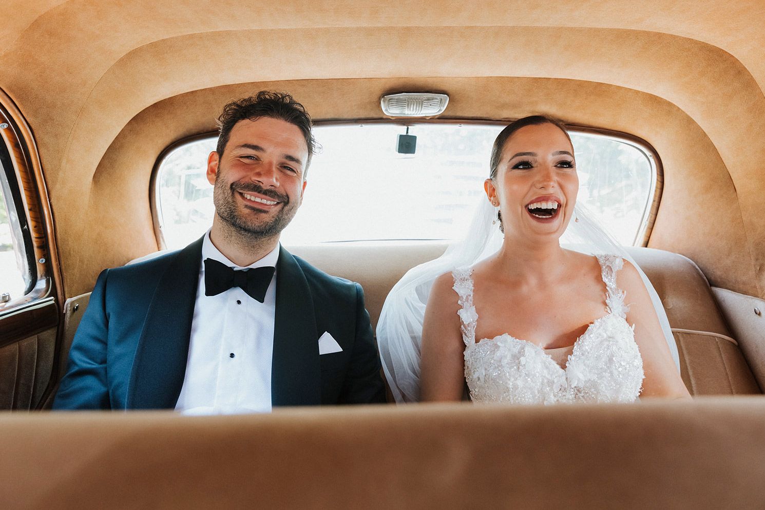 Unscripted joy as a bride and groom laugh heartily in the back seat of a vintage getaway car during their German-Turkish wedding day.