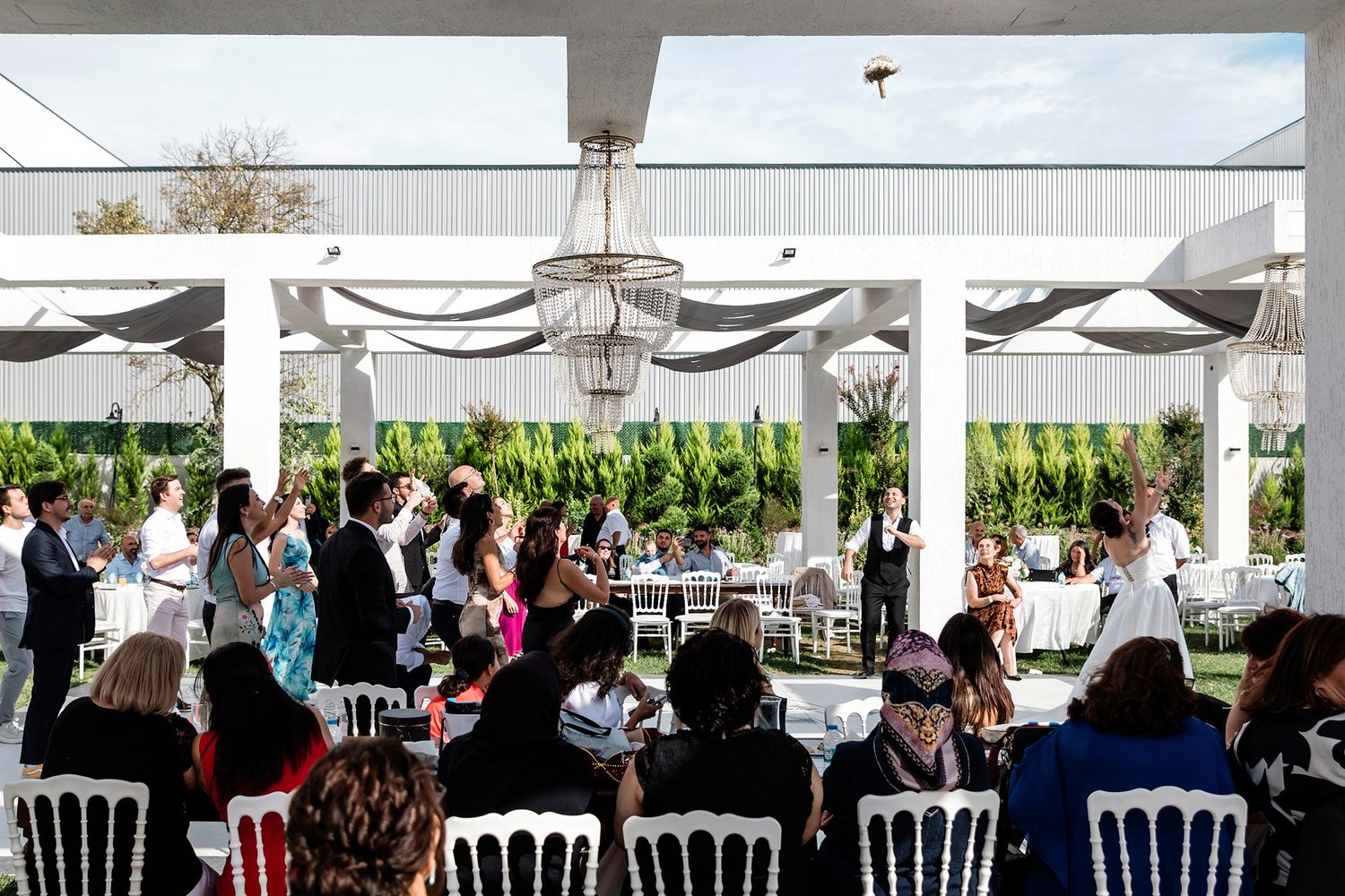 Wide shot of a bride throwing her bouquet high into the air as excited guests cheer during an elegant outdoor garden wedding reception.