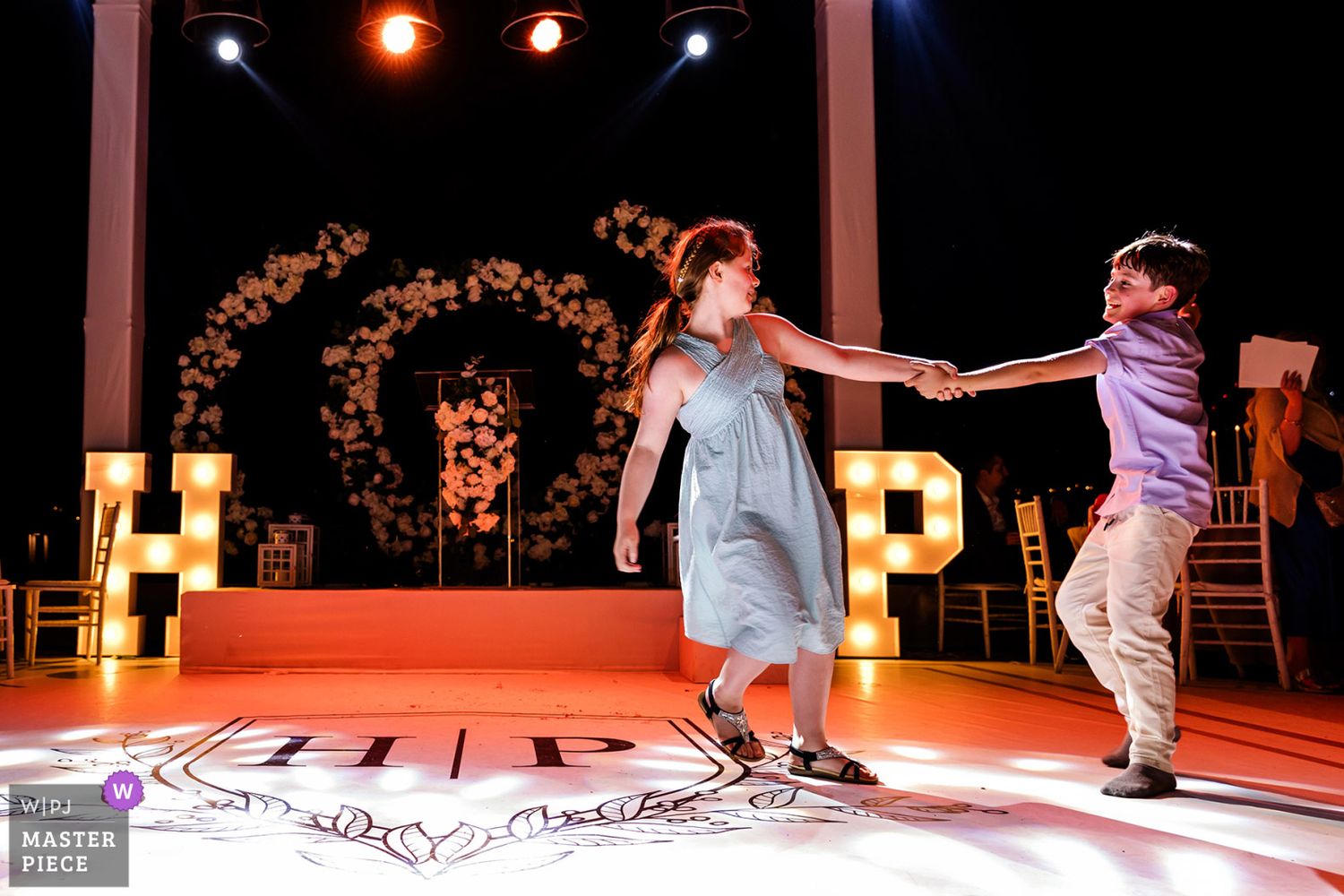 WPJA award-winning candid photo of a young boy and girl holding hands and dancing playfully on an illuminated wedding dance floor.