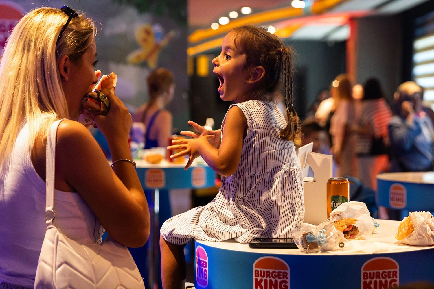 An excited young girl laughing and cheering on a branded table while her mother eats during a high-energy Burger King activation.