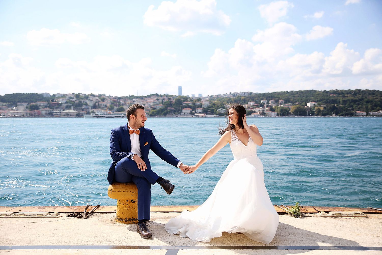 A bride and groom in a blue suit laughing with pure, unscripted joy while sitting on a yellow mooring bollard by the Bosphorus.