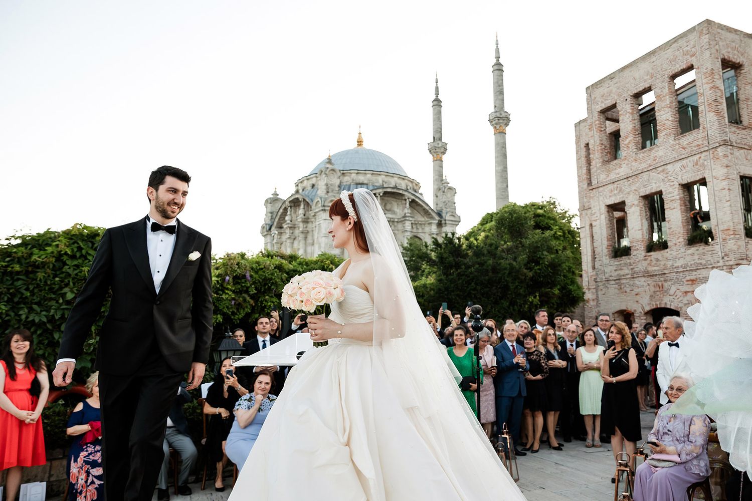 A smiling bride and groom walk down the aisle during their wedding recessional at Esma Sultan, with the Ortaköy Mosque behind them.