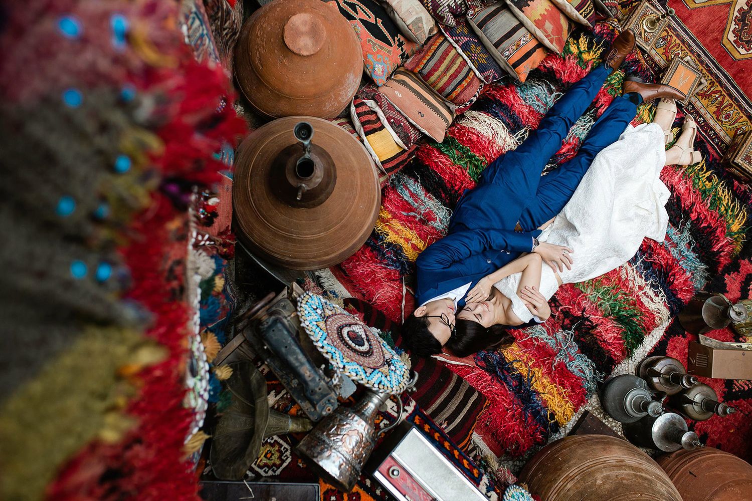 Top-down view of a bride and groom lying intimately on vibrant traditional Turkish rugs surrounded by pottery in Cappadocia.