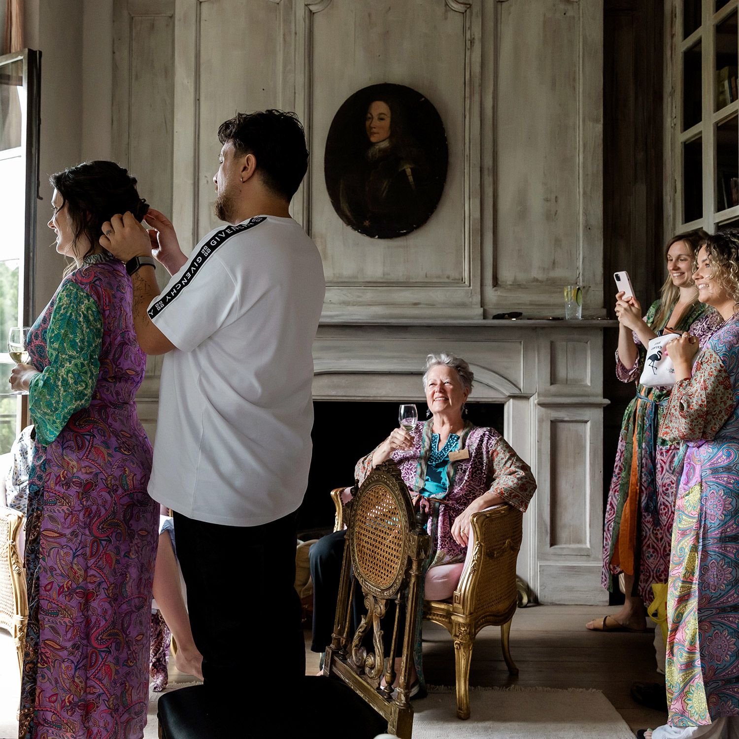 A relaxed bride gets her hair styled while guests drink wine and bridesmaids snap photos in a historic room at Aslı Tunca, Istanbul.