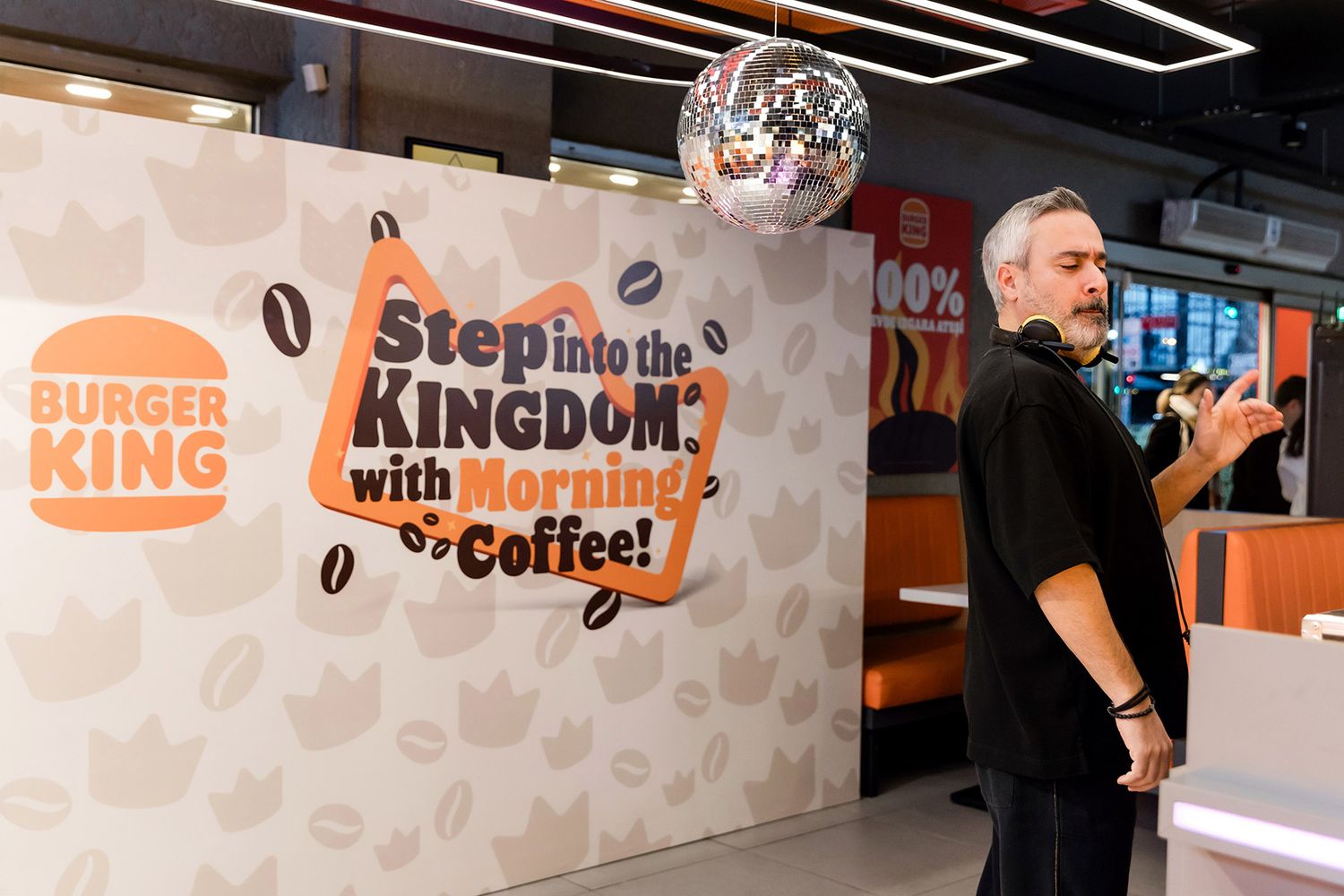 A live DJ performing under a disco ball next to a branded photo backdrop for a lively Burger King morning coffee brand activation.