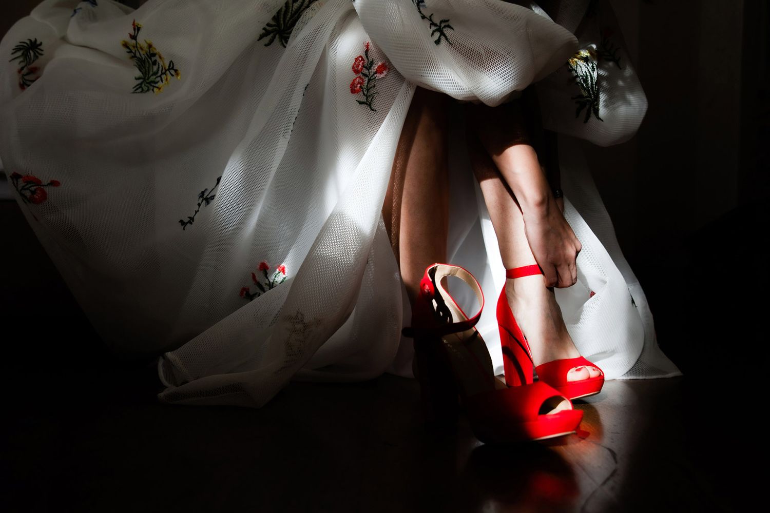 Dramatic documentary detail shot of a bride putting on vibrant red high heels under her floral embroidered wedding dress.