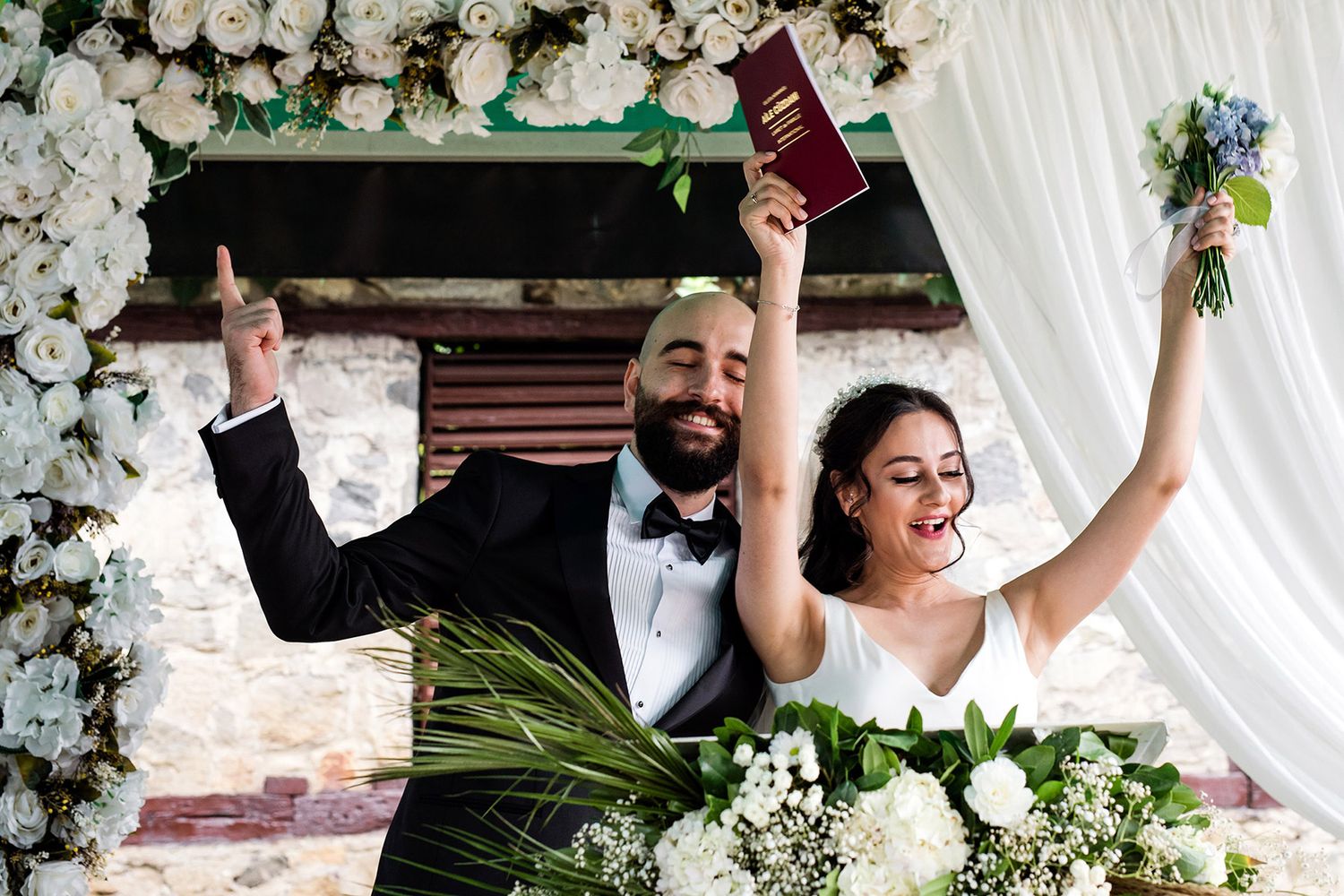 A joyful bride and groom cheer with their hands raised, holding the red Turkish marriage booklet after their ceremony at Bursa Bey Konağı.