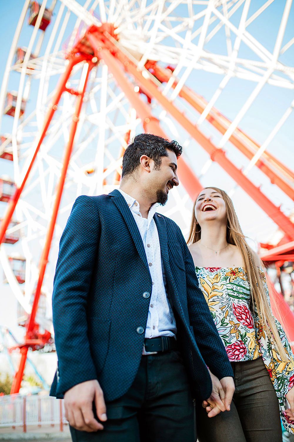 An engaged couple laughs genuinely while holding hands in front of a massive red Ferris wheel during a playful, unscripted shoot in Bursa.