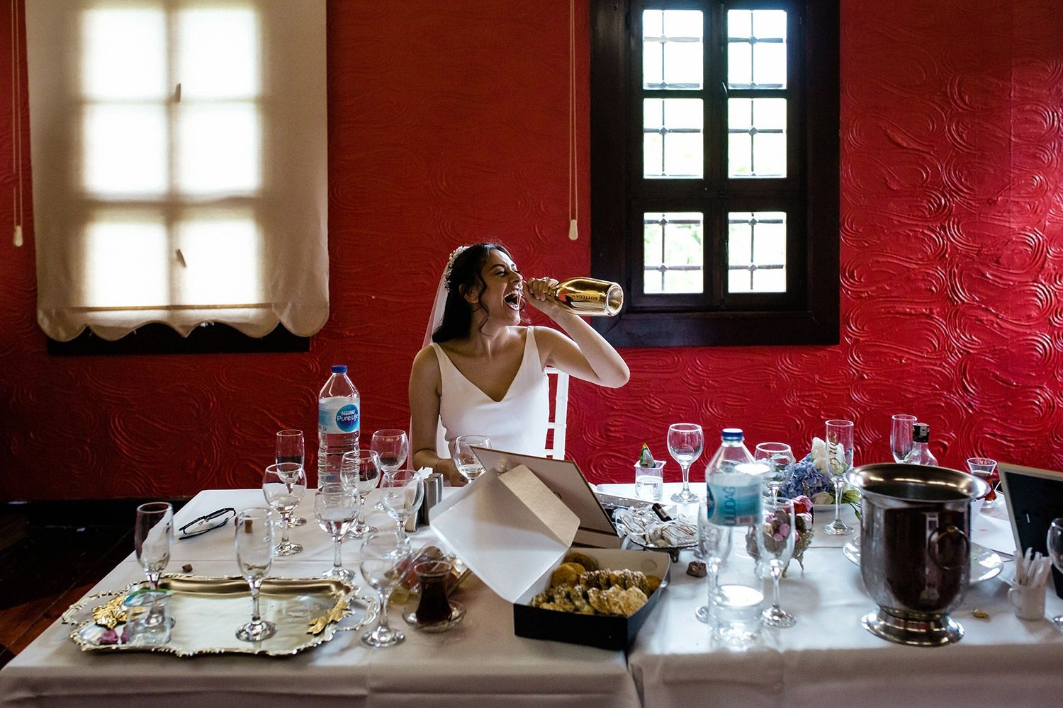 Candid photo of a laughing bride drinking from a gold champagne bottle at a wedding dinner table with a vibrant red wall.