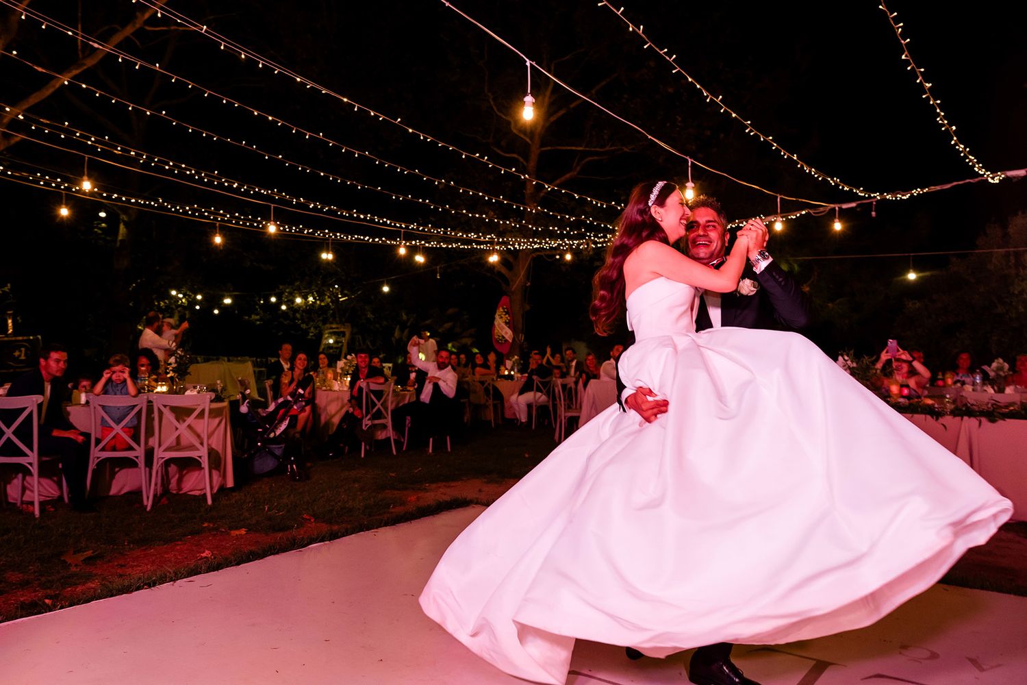 A groom joyfully carries and spins his laughing bride on an outdoor dance floor under string lights during a vibrant Tekirdağ wedding.