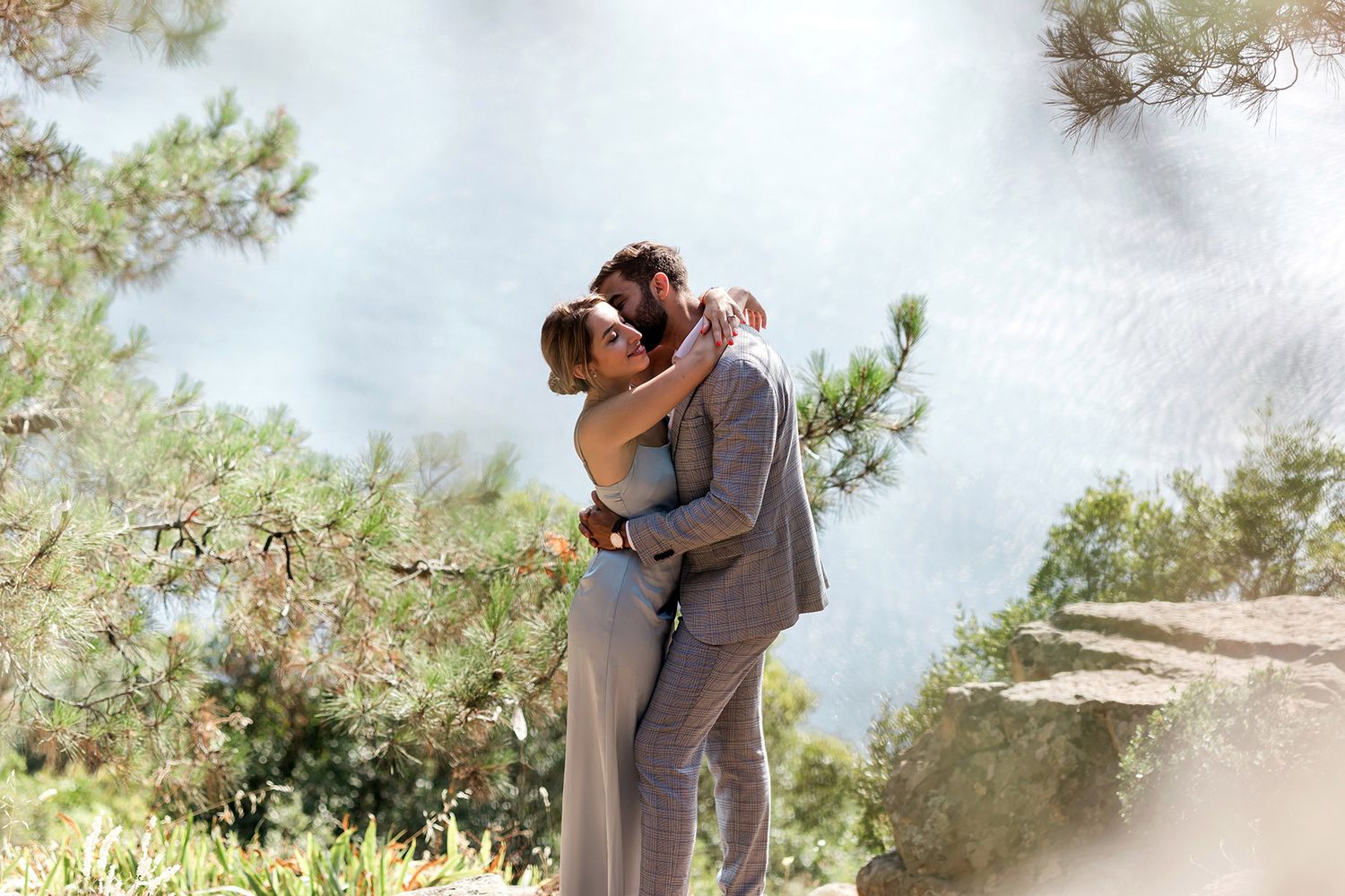 A romantic couple shares a tender, unscripted embrace on a sunlit cliffside surrounded by pine trees on Büyükada Island, Istanbul.