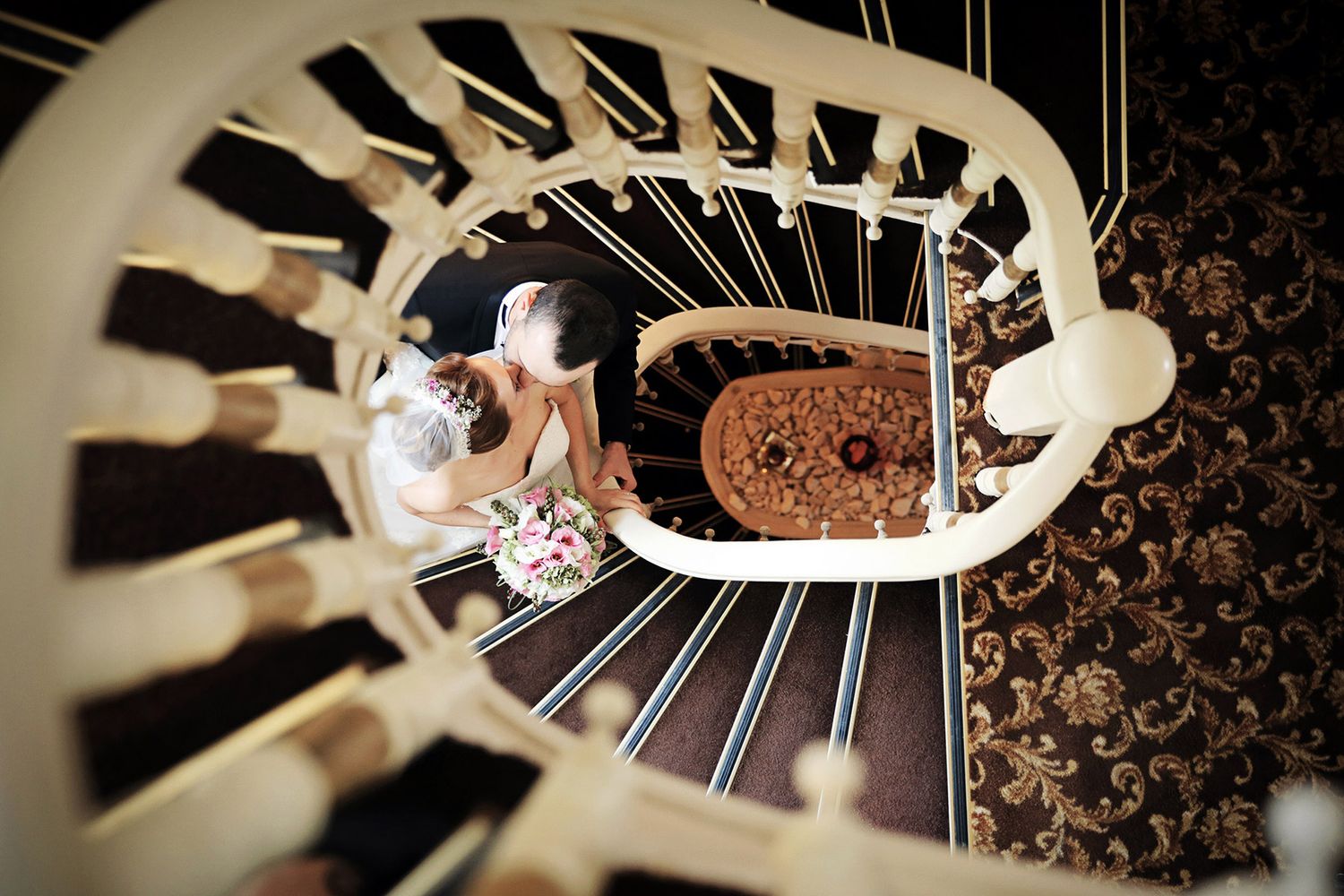 Creative top-down view through a vintage spiral staircase, capturing a bride and groom sharing a romantic kiss at the bottom.
