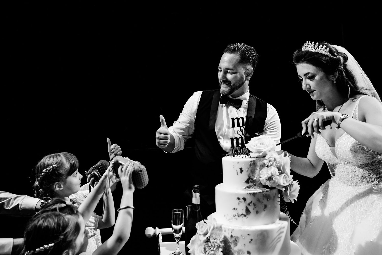 Siyah beyaz düğün fotoğrafı, gelin ve damat pasta keserken çocuklar telefonla fotoğraf çekiyor.