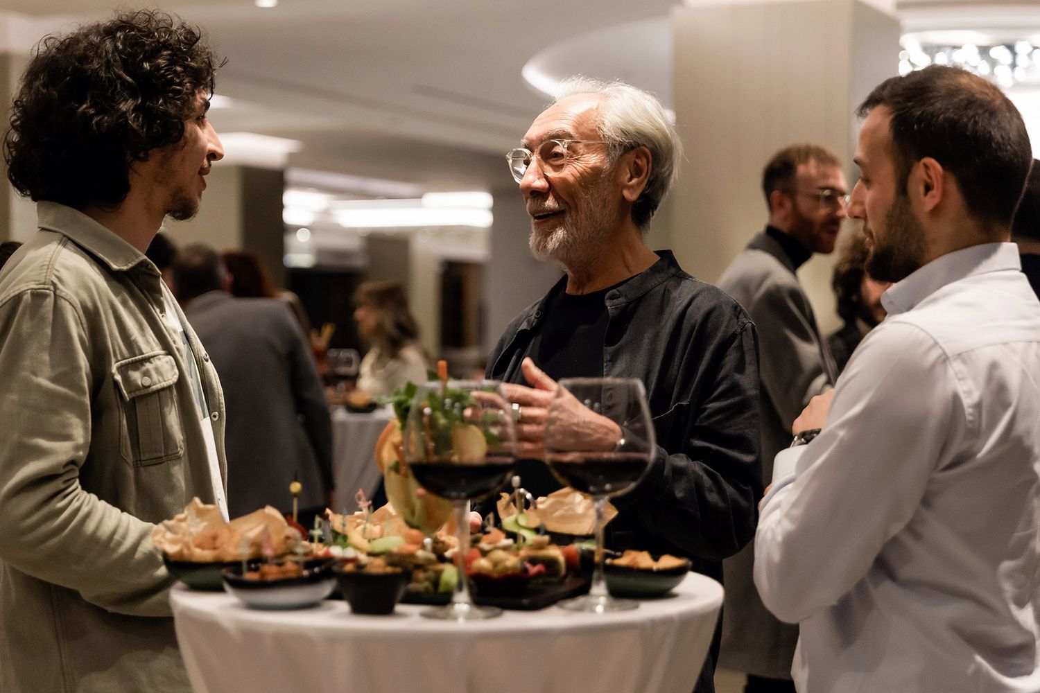 Three professional men engaging in deep conversation around a cocktail table filled with gourmet catering at a VIP corporate reception.