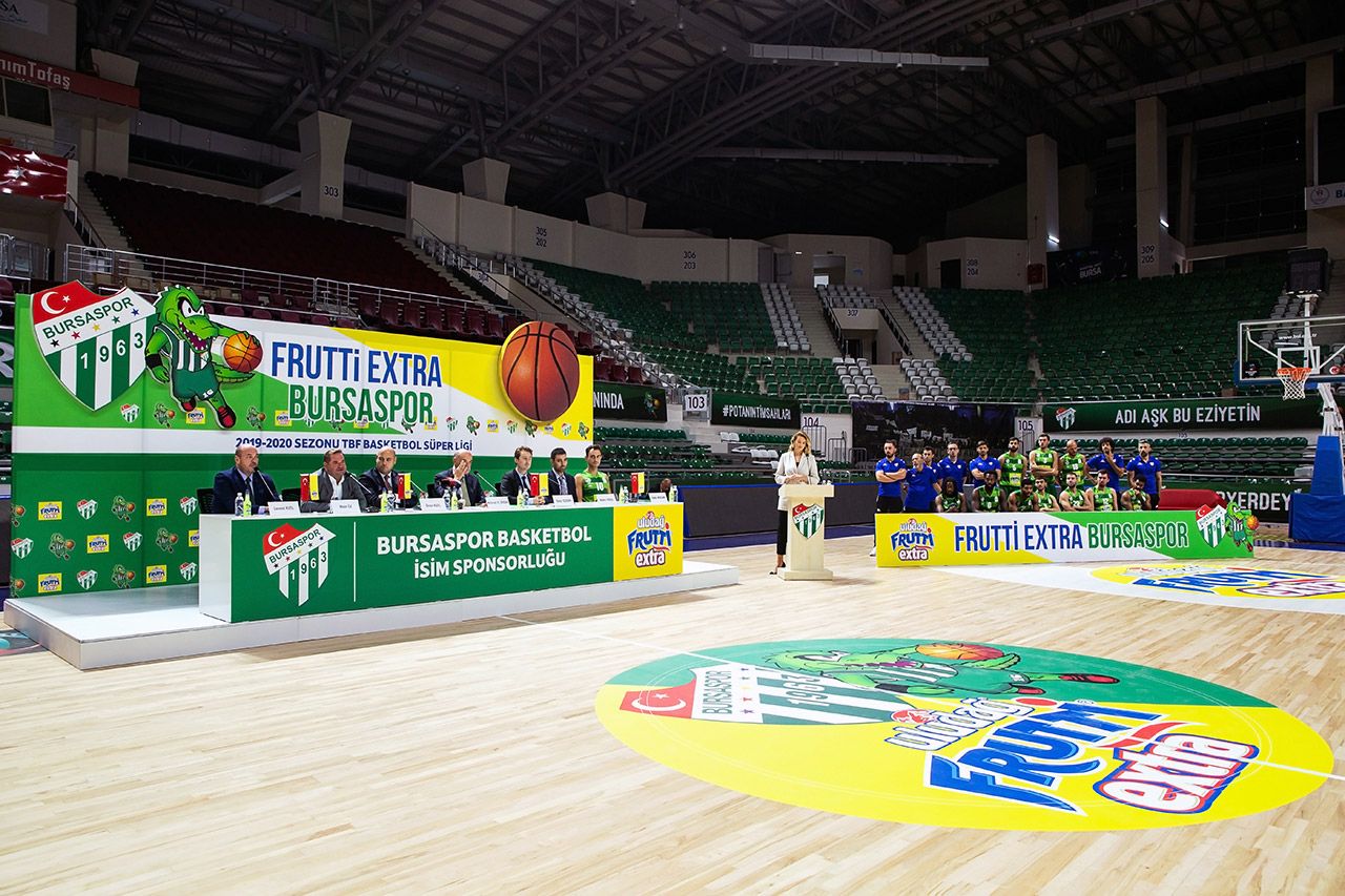 A wide view of a basketball arena showing a corporate sponsorship press conference setup on the court, featuring executives and players.
