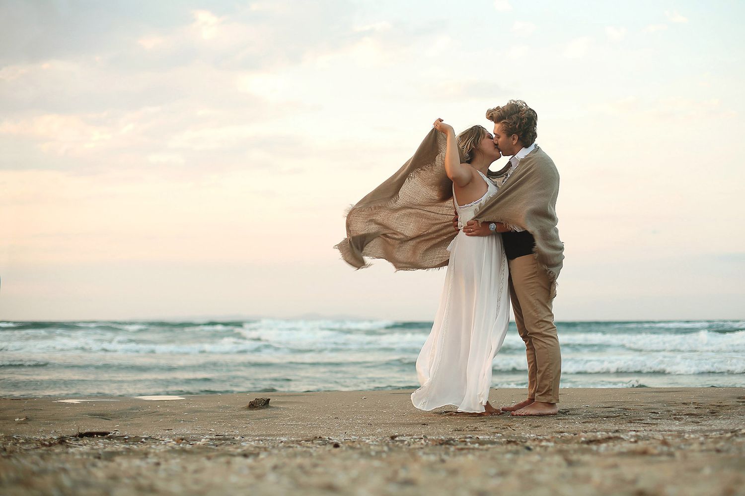 A bride and groom share a romantic kiss while wrapped in a large blanket blowing in the wind on a quiet, overcast beach in Bursa.