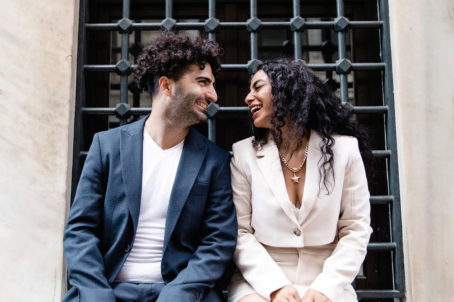 A stylish couple in a suit and white blazer sitting and laughing out loud against a black iron window grate in Bankalar Caddesi, Istanbul.