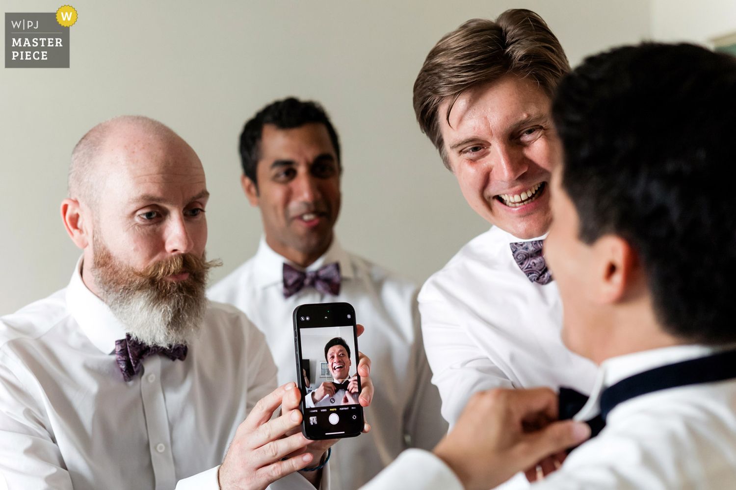 WPJA award-winning candid photo of laughing groomsmen using a smartphone camera as a mirror for the groom to tie his bowtie.