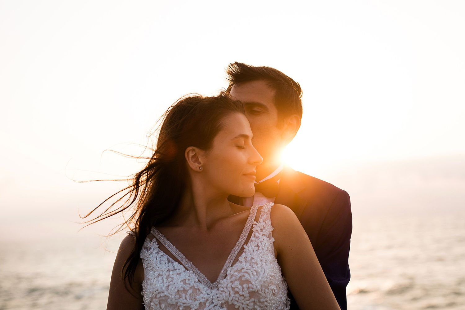 A groom gently kisses his bride's forehead during a romantic, sun-drenched golden hour portrait session with beautiful lens flare.