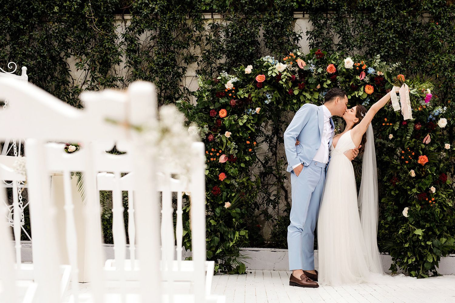 A bride and groom kiss passionately in front of an ornate floral arch, the groom in a light blue suit, framed by white chairs.