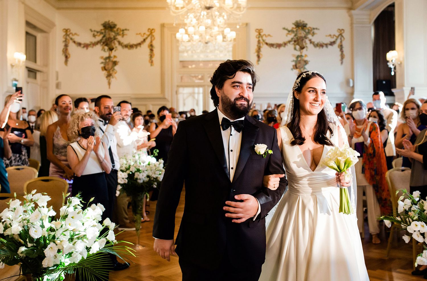 A joyful bride and groom make their grand entrance down the aisle, surrounded by clapping guests at the elegant Pera Palace Hotel.