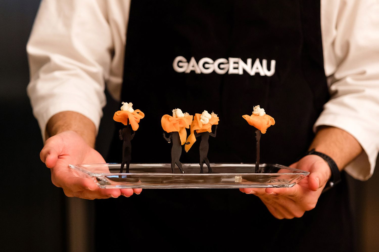 Close-up of a chef in a Gaggenau apron holding a glass tray of highly creative, branded gourmet appetizers.