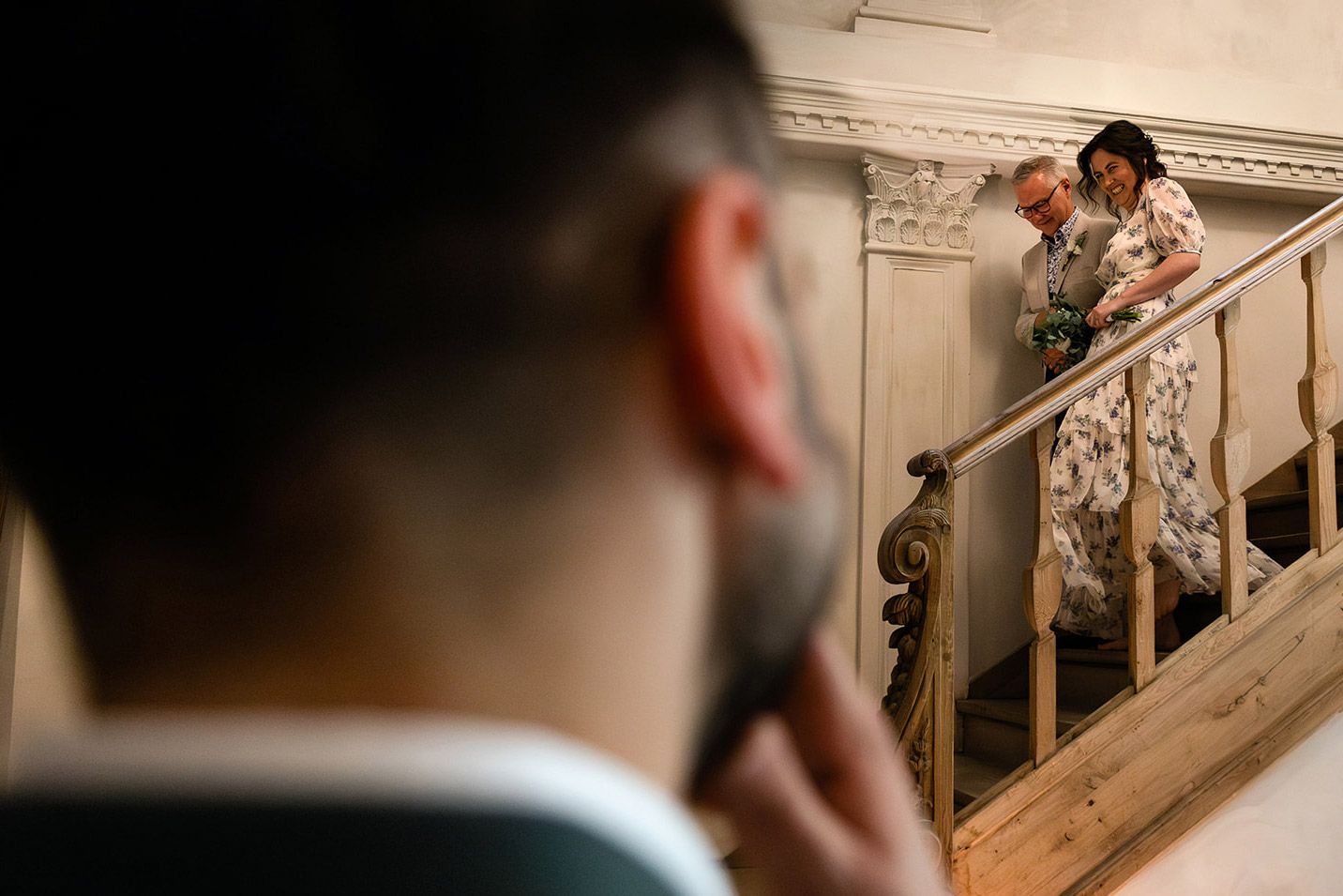 The groom watches in the foreground as his smiling bride walks down a vintage wooden staircase during an Aslı Tunca boutique wedding.