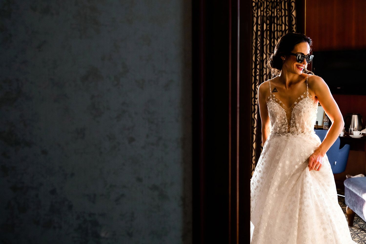 A cool, smiling bride wears black sunglasses and her wedding dress in a moody, dramatic doorway during her unscripted Istanbul prep.